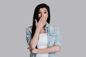 No way Surprised attractive young woman in jeans wear covering mouth with hand and looking at camera while standing against grey background photo