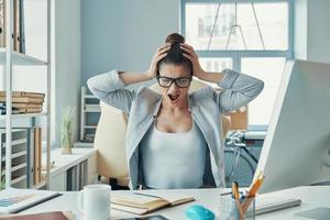 mujer joven estresada con ropa informal inteligente manteniendo la cabeza en las manos y gritando mientras está sentada en la oficina foto