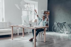 Handsome young man throwing paper in air and smiling while sitting at his working place in creative office photo