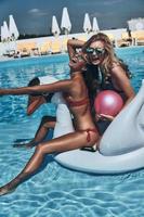 dos hermosas mujeres jóvenes en bikini riéndose mientras flotan en un gran cisne inflable foto