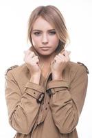 Feeling comfortable and trendy. Attractive young woman in coat adjusting her collar and looking at camera while standing against white background photo