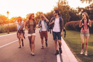 Enjoying summer hike. Group of young people with backpacks walking together by the road and looking happy photo