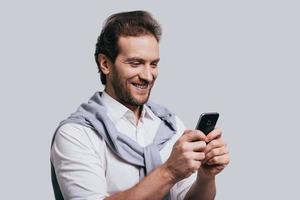 Texting to friend. Beautiful young man in smart casual clothes typing a message and smiling while standing against grey background photo