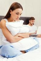 Feeling lonely near him. Depressed young woman embracing pillow and looking down while sitting on bed with young man working on laptop in the background photo