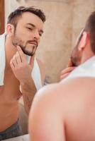 To shave or not to shave. Handsome young man touching his face and smiling while standing in front of the mirror photo