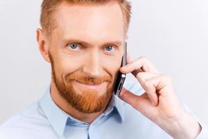 buenas noticias. primer plano de un hombre barbudo hablando por teléfono móvil y sonriendo mientras está de pie contra el fondo blanco foto