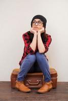 Feeling bored.  Beautiful young woman in headwear holding hands on chin and looking away while sitting on suitcase photo