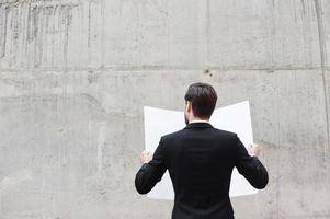 Concentration at work. Rear view of young man examining blueprint while standing outdoors and against the concrete wall photo