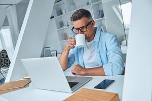 Confident mature man working on laptop and drinking coffee while sitting at the office desk photo