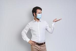 Confident young man in white shirt wearing protective face mask and gesturing while standing against gray background photo