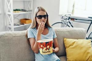 Beautiful young woman watching TV in 3-D glasses and eating popcorn while relaxing on the sofa at home photo