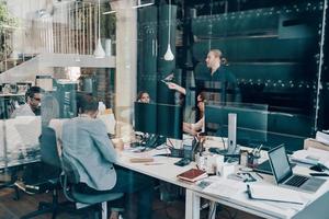Group of young modern people in smart casual wear communicating and using modern technologies while working in the office photo