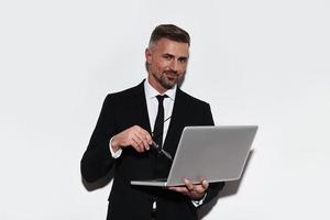 Always ready to help. Handsome young man in full suit using laptop and looking at camera with smile while standing against white background photo