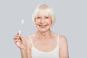 Dental health. Beautiful senior woman holding toothbrush and smiling while standing against grey background photo