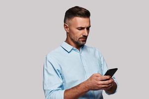 Very busy. Good looking young man in smart casual wear using his smart phone and smiling while standing against grey background photo