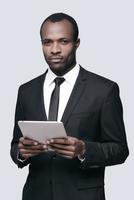 Confident and handsome. Young African man in formalwear working on digital tablet while standing against grey background photo