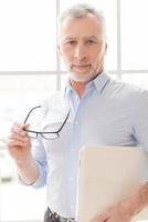 Trust to professionals. Confident grey hair senior man in shirt holding laptop and looking at camera while standing in front of the window photo