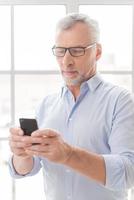 Typing business message. Serious grey hair senior man in shirt holding mobile phone and looking at it while standing in front of the window photo