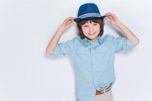 disfrutando de su estilo. niño feliz ajustando su sombrero y sonriendo mientras está de pie contra el fondo blanco foto