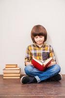 leyendo su libro favorito. niño leyendo un libro mientras se apoya en la pared con una pila de libros cerca de él foto