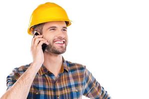 Happy with work done. Happy young male carpenter in hardhat talking on mobile phone and smiling while standing against white background photo