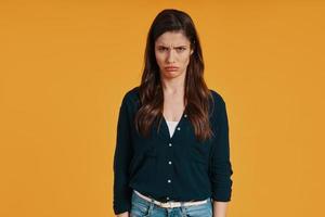 Sad young woman in casual clothing making a face while standing against yellow background photo