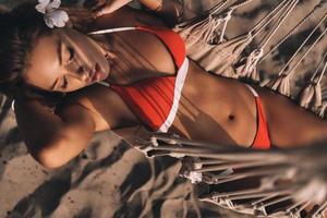 Pure sensuality. Top view of attractive young woman in swimwear keeping hands behind head while lying down in hammock on the beach photo