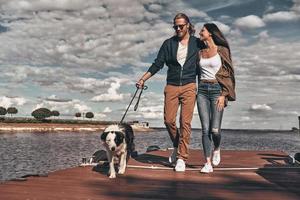 Enjoying nice weekend. Full length of beautiful young couple walking with their dog on the river shore while spending time outdoors photo