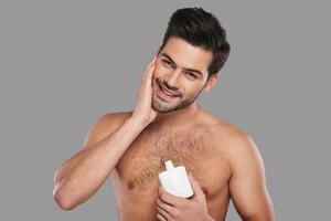 Feeling so fresh. Handsome young man applying aftershave lotion and smiling while standing against grey background photo