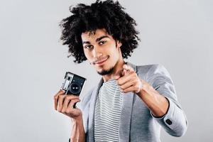 ¿Puedo tomar una foto de usted, un joven africano guapo sosteniendo una cámara de estilo retro y apuntándolo mientras está de pie contra un fondo gris?