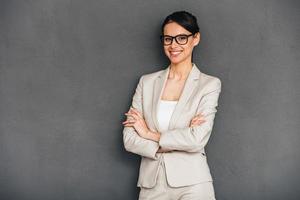 Cheerful and confidant. Cheerful young businesswoman in glasses keeping arms crossed and looking at camera with smile while standing against grey background photo