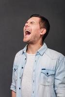 So furious Young handsome man keeping mouth open and eyes closed while standing against grey background photo