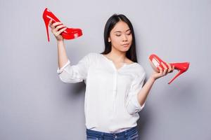 Eternal dilemma. Beautiful young Asian woman holding shoes of different colors while standing against grey background photo