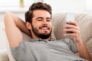 Happy to be at home. Smiling bearded man holding mobile phone and looking on it while laying on sofa photo