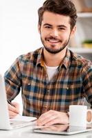 frente a la satisfacción laboral. un joven sonriente escribiendo en un bloc de notas y mirando la cámara mientras se sienta en su lugar de trabajo foto