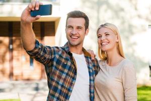 Selfie for their family album. Cheerful young couple bonding to each other while making selfie by smart phone while standing against their new house photo