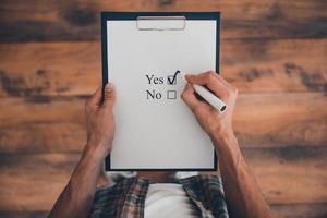 Making a right choice. Top view of man making a check mark in clipboard while standing on the wooden floor photo