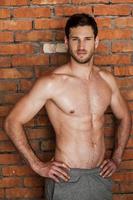 Strong handsome. Handsome young shirtless man holding hands on hips and looking at camera while standing against brick wall photo
