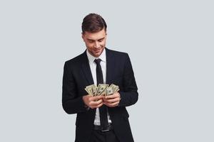 Always in profit. Good looking young man in full suit counting money and smiling while standing against grey background photo