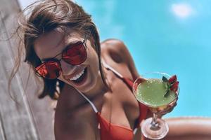 felicidad verdadera. vista superior de una atractiva joven en traje de baño sosteniendo un vaso y sonriendo mientras se sienta junto a la piscina al aire libre foto