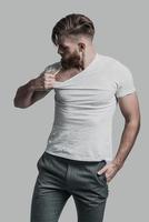 Carefree look. Young man pulling his T-shirt while standing against grey background photo
