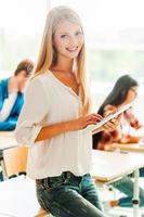 Studying in a digital way. Beautiful young woman holding digital tablet and looking at camera while leaning at the desk photo