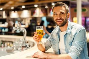 Tiempo de cerveza. joven alegre sosteniendo un vaso de cerveza y mirando a la cámara mientras se sienta en el mostrador del bar foto