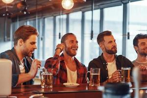 grupo de jóvenes felices con ropa informal disfrutando de la cerveza mientras se sientan en el mostrador del bar en el pub foto