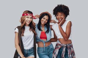 tres atractivas mujeres jóvenes con estilo mirando a la cámara y sonriendo mientras están de pie contra el fondo gris foto