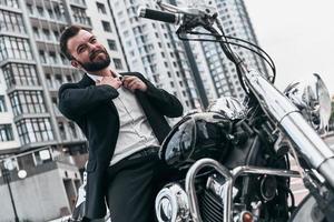 Tired of being serious. Good looking young man in full suit taking off his necktie and smiling while sitting on the motorbike outdoors photo