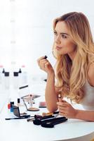 Time for lipstick. Side view of beautiful young woman applying lipstick and looking at her reflection in mirror while sitting at the dressing table photo