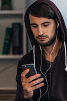 triste canción. joven pensativo con camisa encapuchada usando su teléfono inteligente mientras está de pie en casa foto