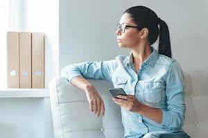 Waiting for important message. Beautiful young woman in glasses holding smart phone and looking at the window while sitting on the couch in office photo