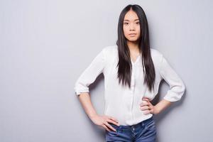 Confident beauty. Beautiful young Asian woman looking at camera and holding hands on hips while standing against grey background photo